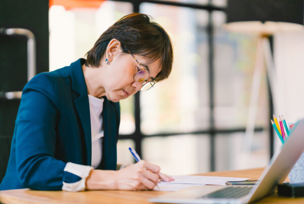 Middle-aged business owner making notes on paper with laptop in front of her