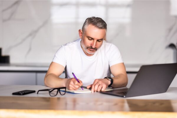 A man with a laptop writes in a notepad